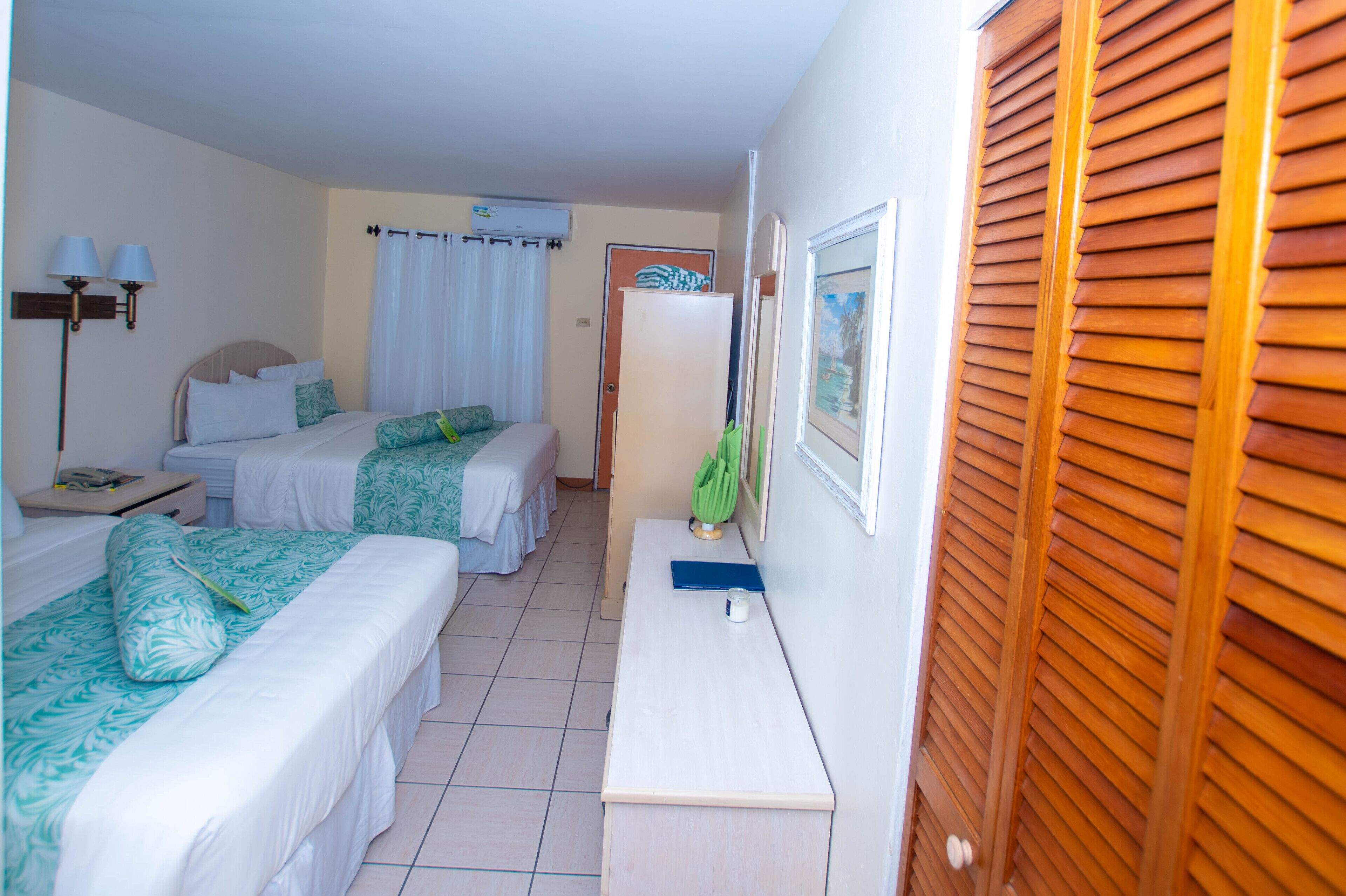 A hotel room with two beds, a desk, a chair, a lamp, and a window with wooden shutters.