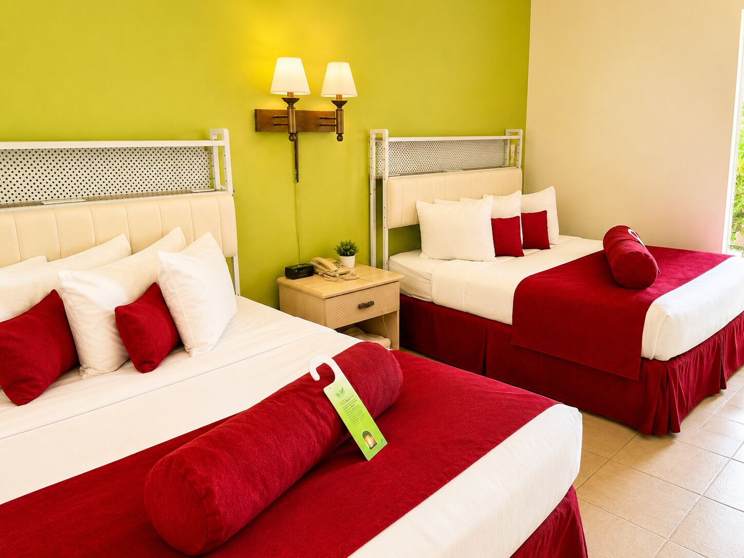 A hotel room with two beds, each with red bedding and white pillows, a nightstand with a lamp, and a green wall.