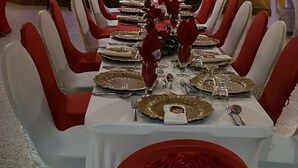 Une longue table à manger, dressée pour un événement formel, avec des chaises blanches et rouges, de la vaisselle dorée et rouge, et un centre de table floral.