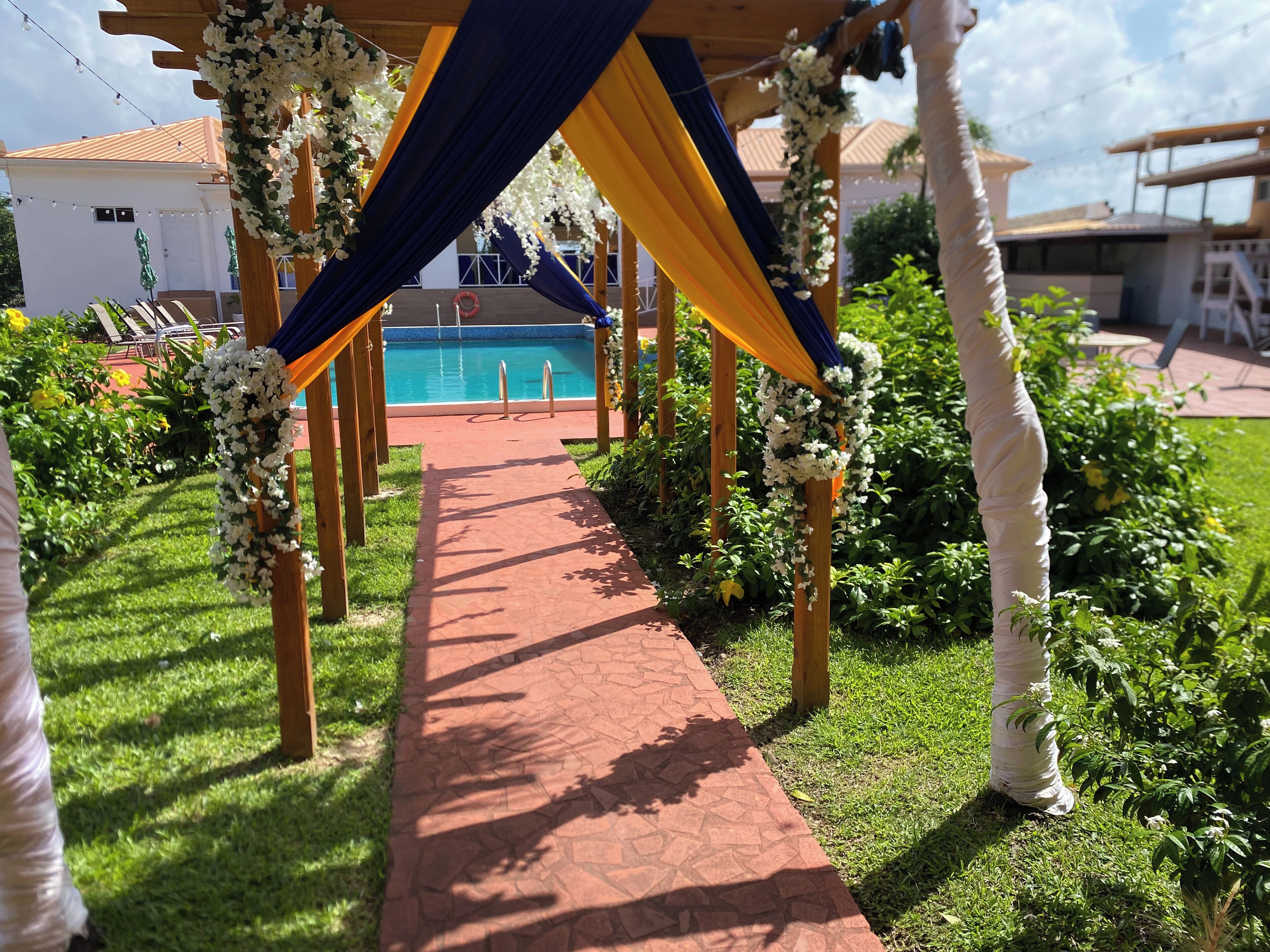 Um caminho leva a uma área de piscina, rodeada por vegetação e uma pérgula de madeira com decorações florais.