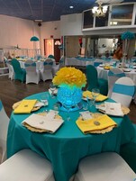 Un salón de banquetes con mesas redondas cubiertas con manteles color turquesa, sillas blancas con cintas turquesa y una pieza decorativa central.