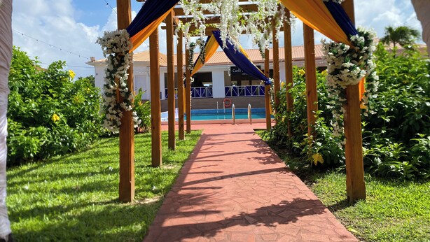 Un chemin mène à un espace piscine, entouré de verdure et agrémenté d’une pergola en bois décorée de fleurs.