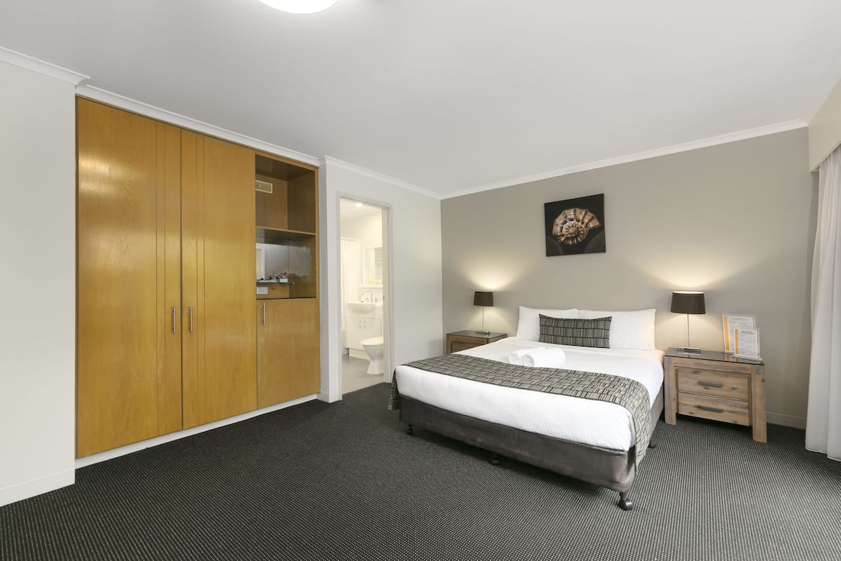Een hotelkamer met een grote kast, een bed met een bedsprei met dessin, twee nachtkastjes met lampen, en een kunstwerk aan de wand.