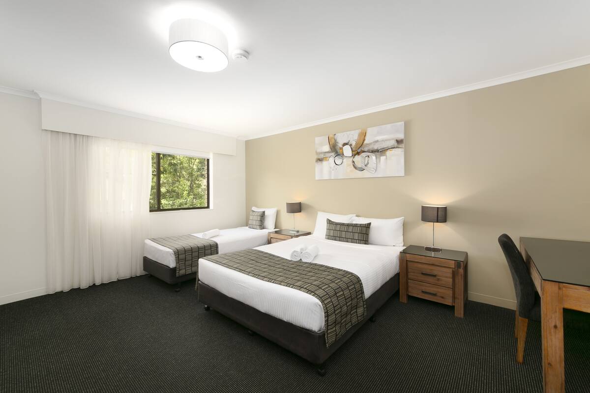 Een hotelkamer met twee bedden, een bureau en een stoel. Er is een raam met gordijnen, een schilderij aan de muur en een lampje op het nachtkastje.