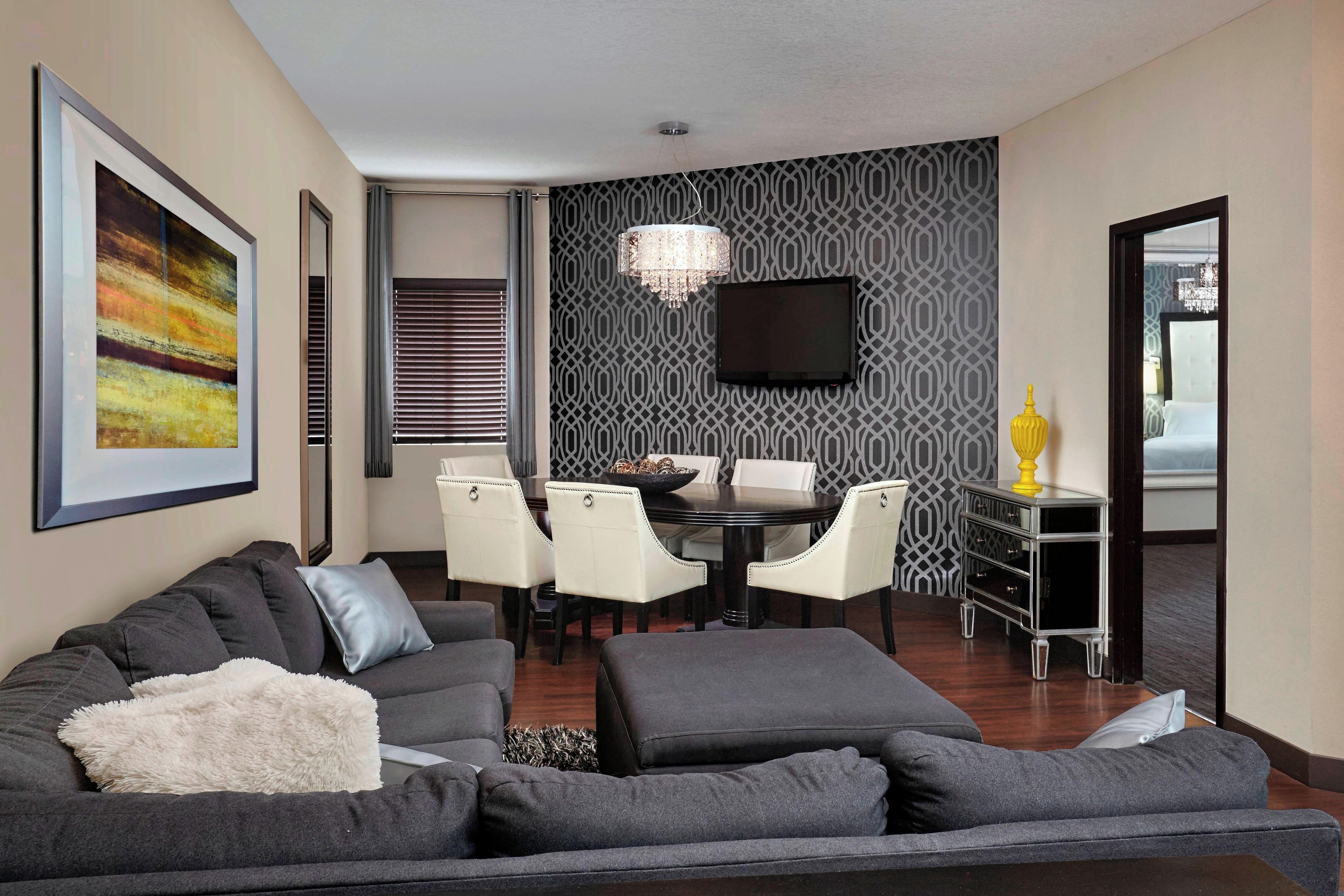 A modern living room with a grey sofa, a dining area with a chandelier, and a TV mounted on a patterned wall.