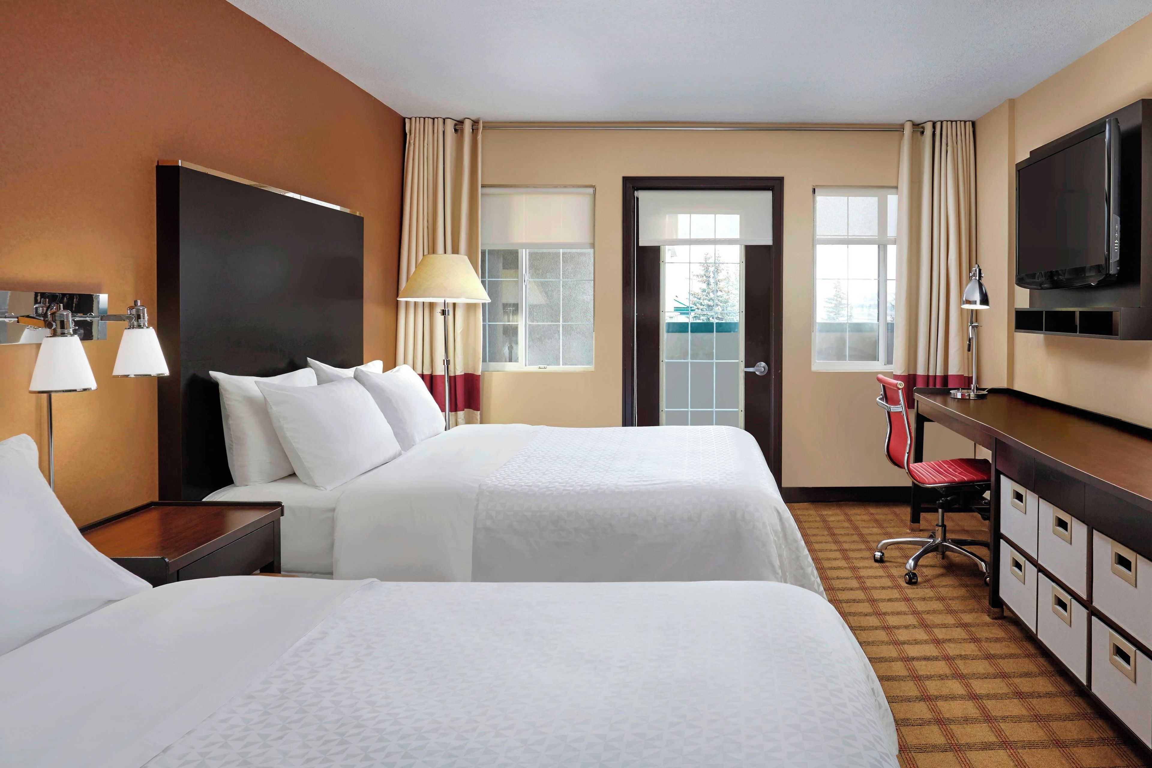 A hotel room with two beds, a desk, a television, and a view of the outdoors.