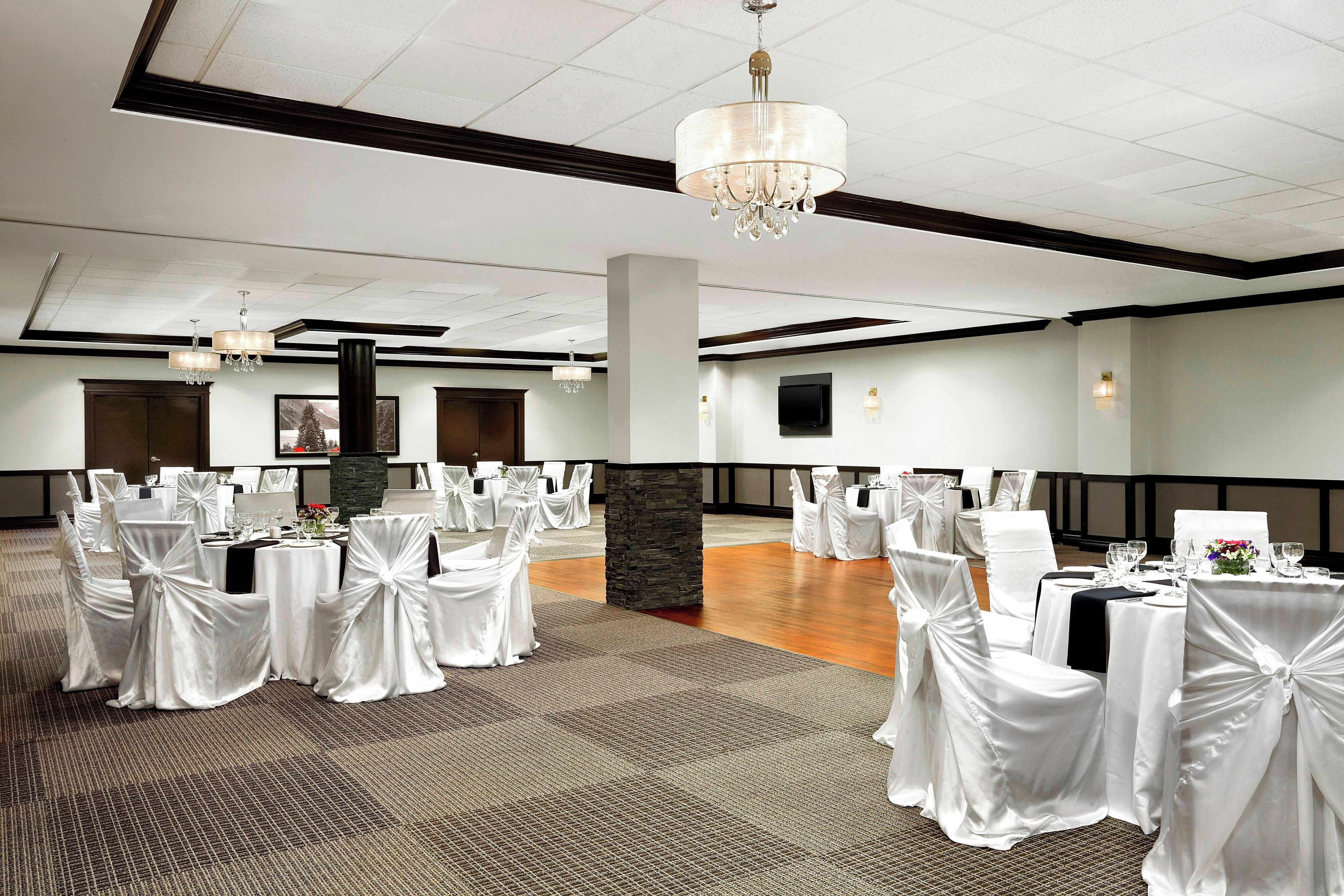 Un salón de banquetes con mesas redondas, sillas blancas, candelabros y un pilar de piedra.