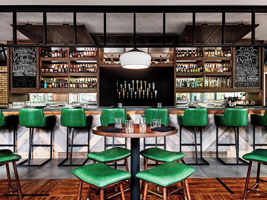 A bar area with green bar stools, a central round table, and shelves stocked with liquor bottles.