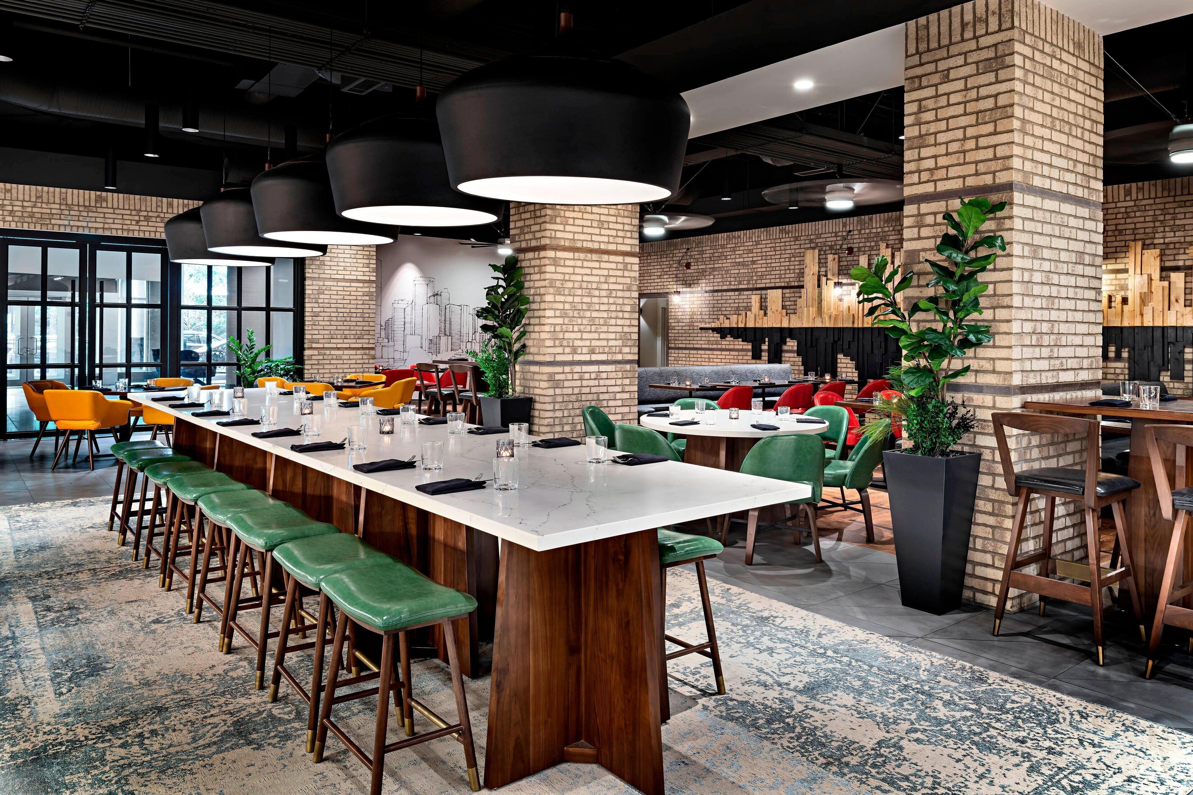 A modern dining area with a long communal table, green bar stools, and red chairs.