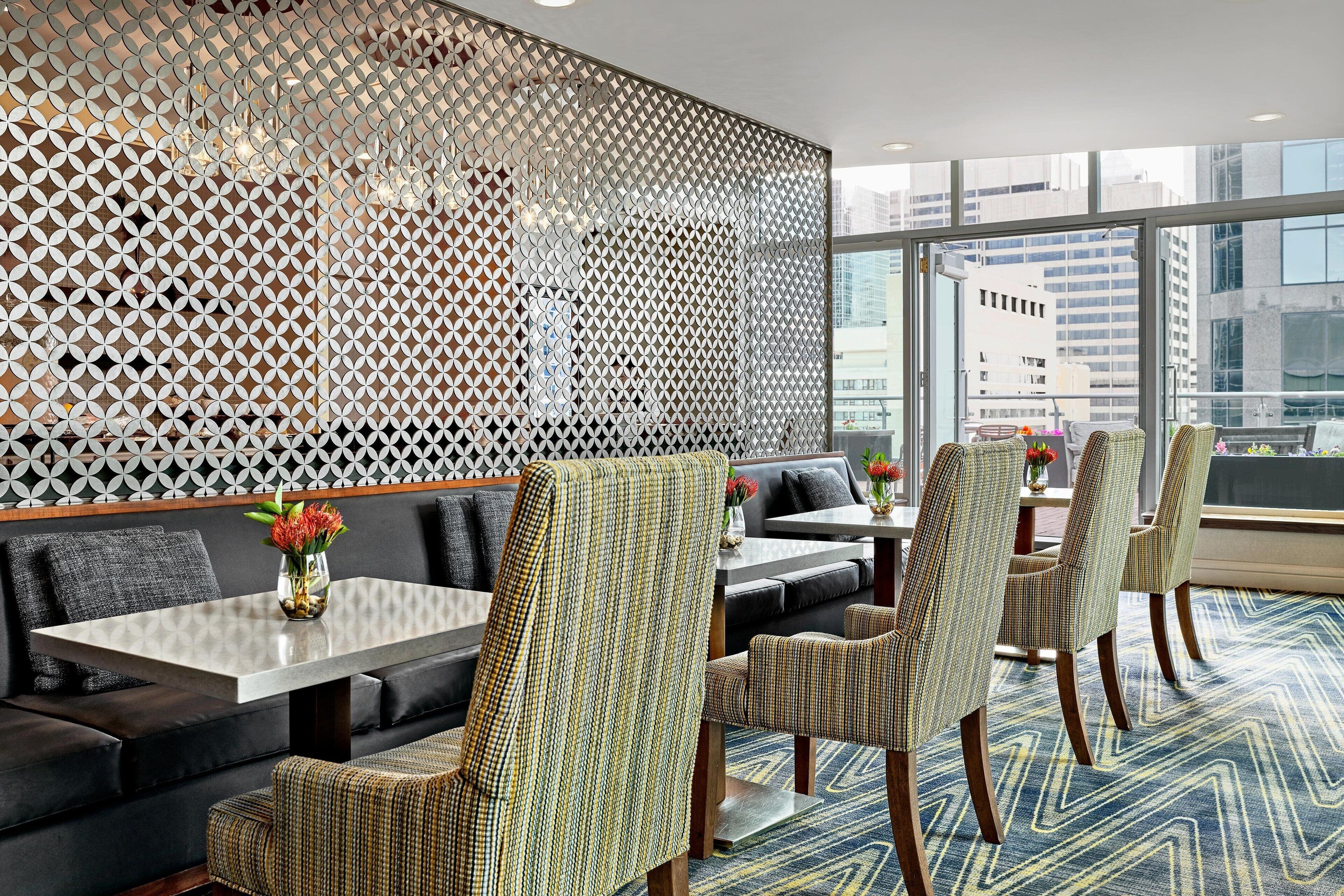 Un comedor moderno con un revestimiento de pared estampado con motivos geométricos, asientos confortables y vistas de la ciudad a través de grandes ventanales.