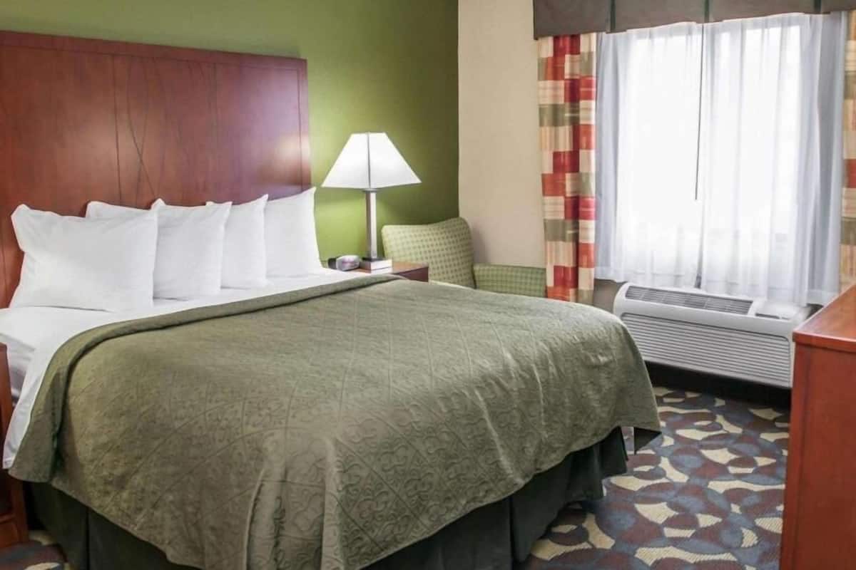 A hotel room with a bed, a nightstand, a lamp, a window with curtains, and a wall heater.