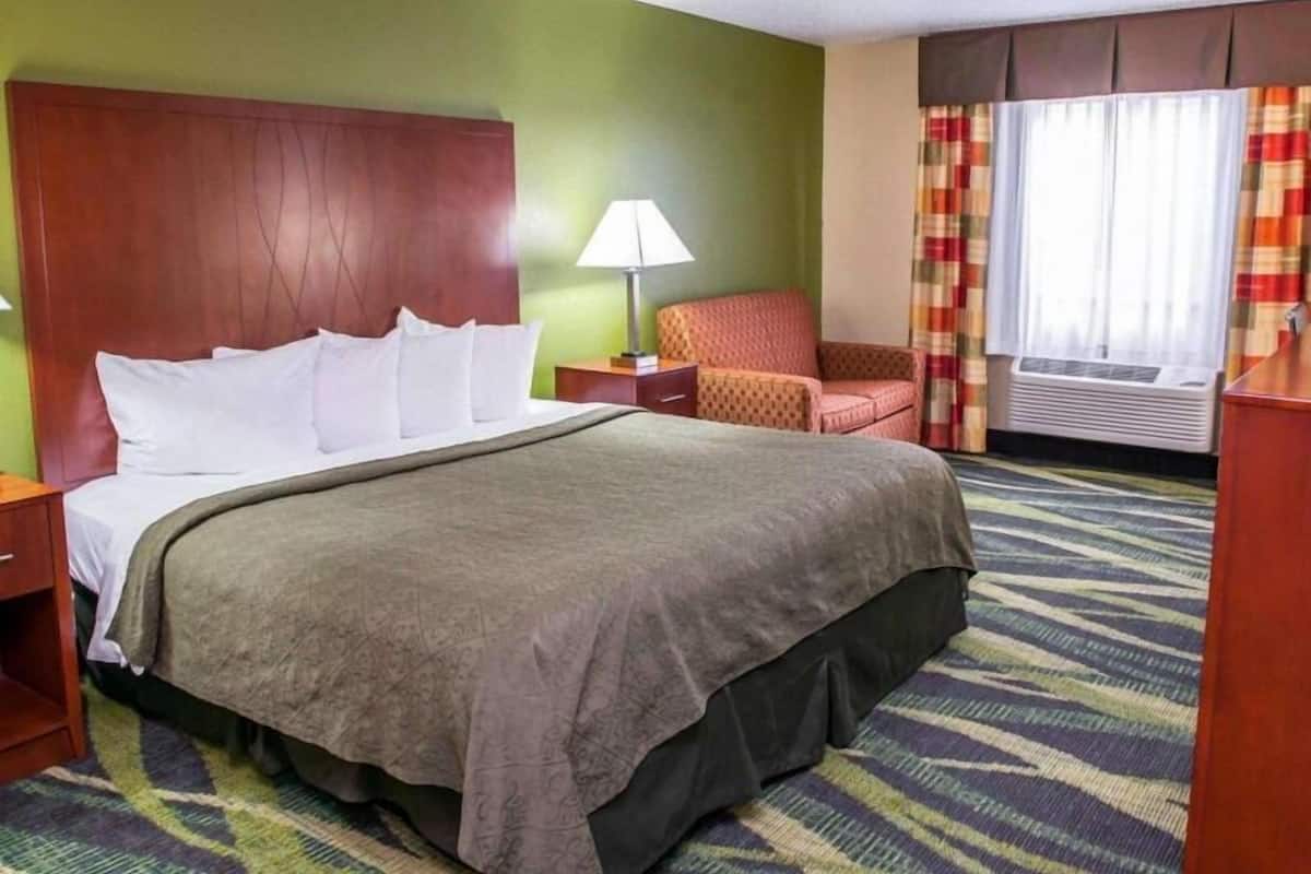 A hotel room with a bed, a chair, a lamp, and an air conditioning unit.