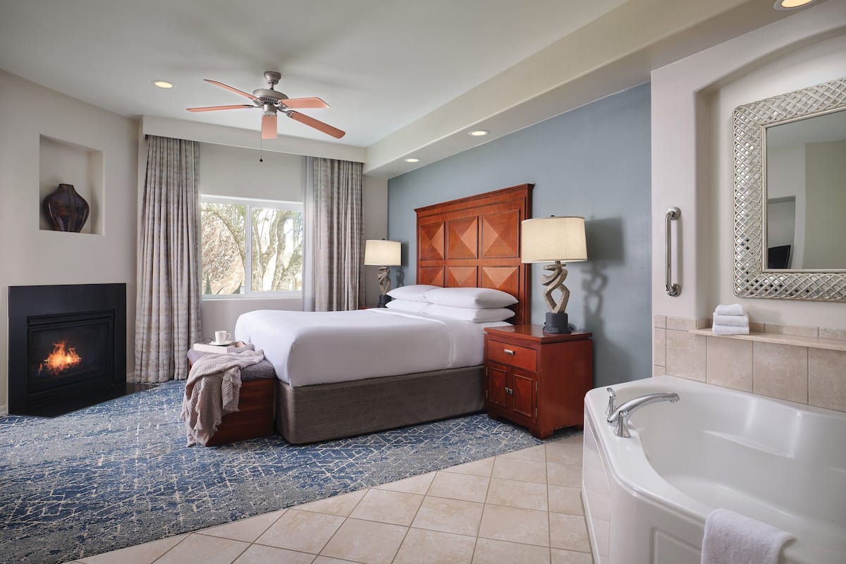 A hotel room with a bed, a fireplace, a large window with curtains, a ceiling fan, a bathroom with a bathtub, and a mirror with a decorative frame.