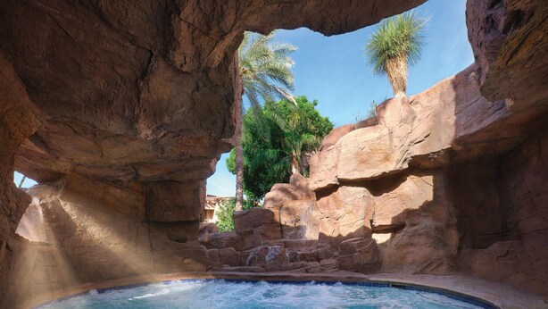 A hot tub set in a natural rock formation with a clear view of palm trees and a waterfall. - Sheraton Desert Oasis Villas, Scottsdale (Scottsdale)