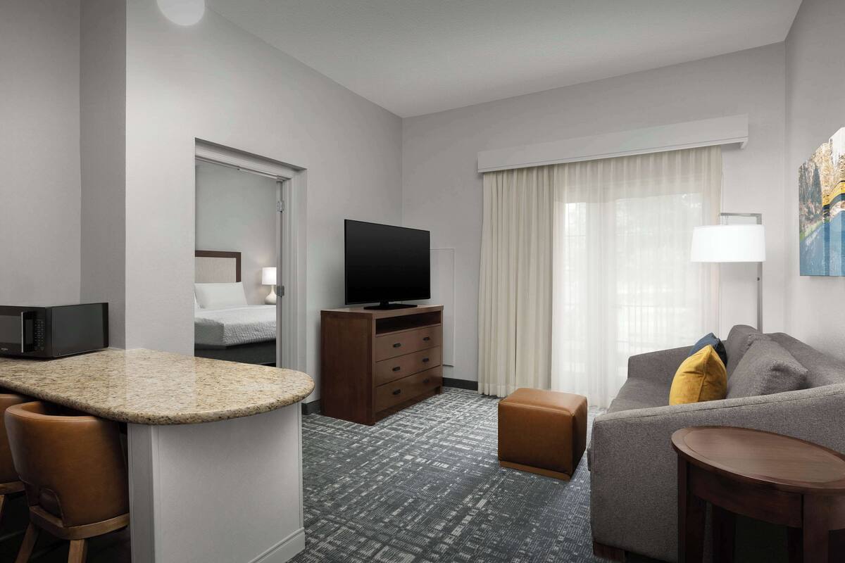 A hotel room with a kitchenette, a flat-screen TV, a sofa, a small table, and a large window with curtains.