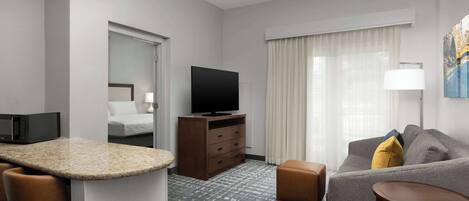 A hotel room with a kitchenette, a flat-screen TV, a sofa, a small table, and a large window with curtains.