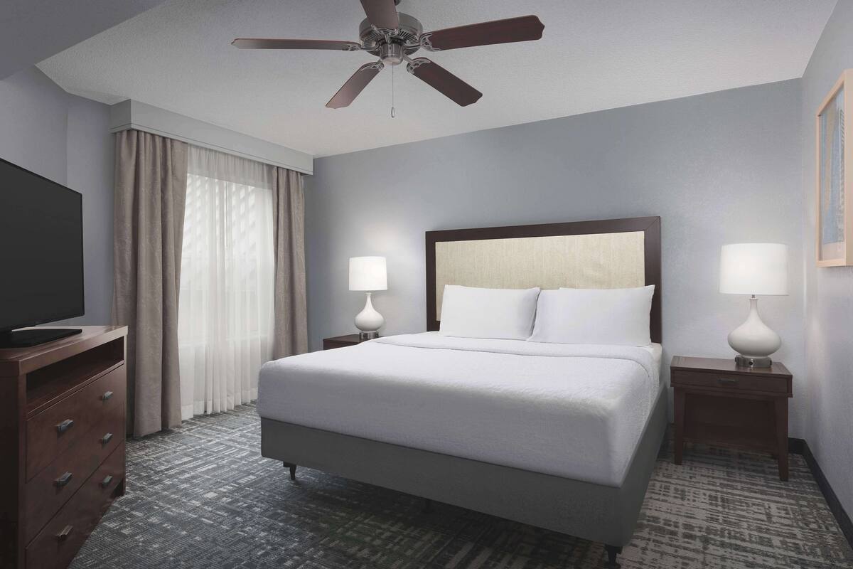 A hotel room with a large bed, two bedside tables with lamps, a ceiling fan, a TV on a wooden dresser, and a window with curtains.