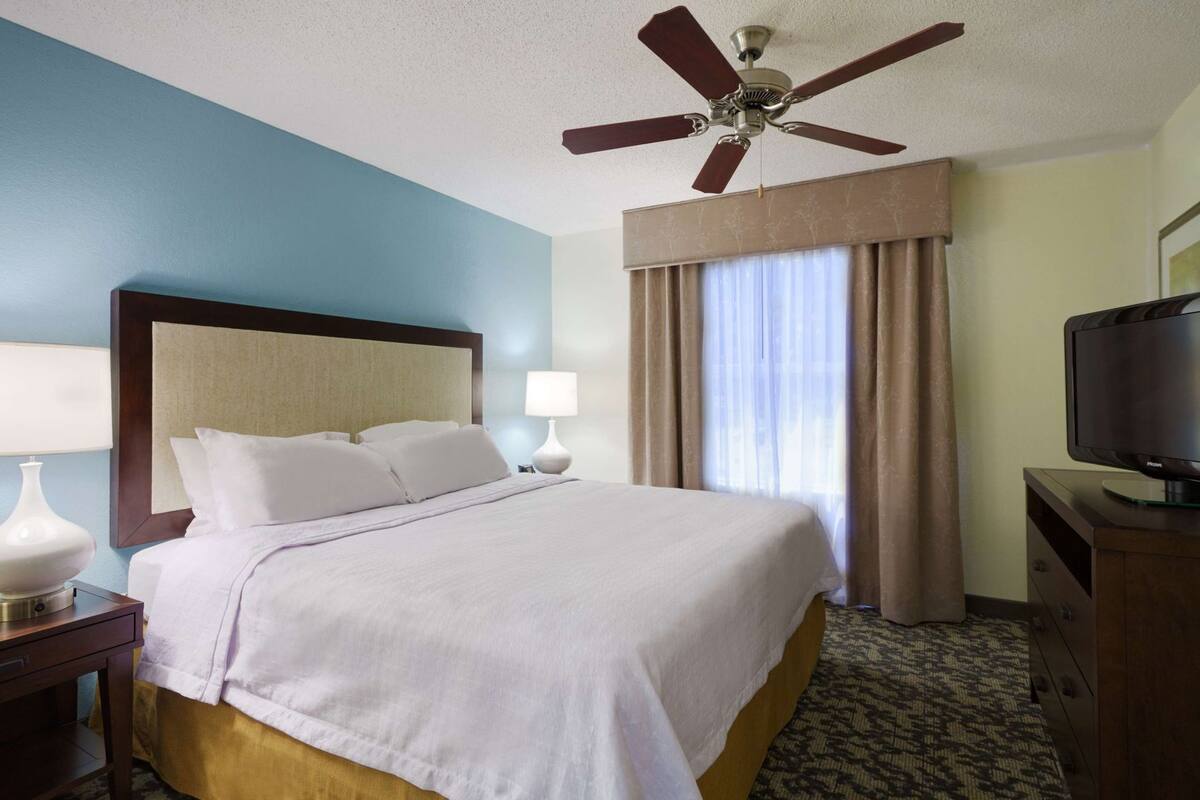 A hotel room with a bed, a ceiling fan, a television, a nightstand, and a window with curtains.