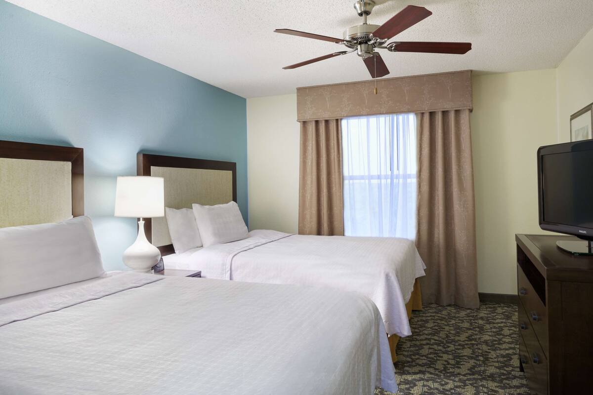 A hotel room with two beds, a ceiling fan, a television, and a window with curtains.