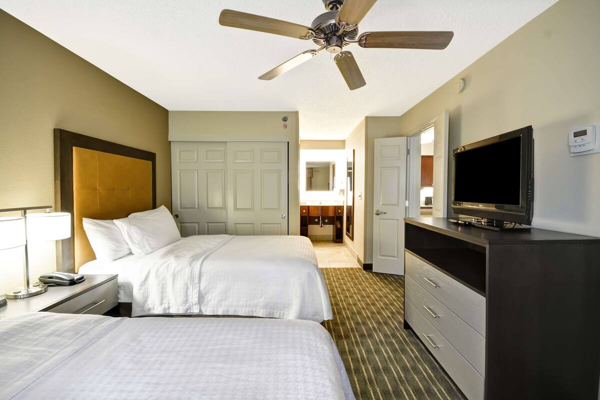 A hotel room with two beds, a ceiling fan, a TV, a desk with a lamp, and a dresser.