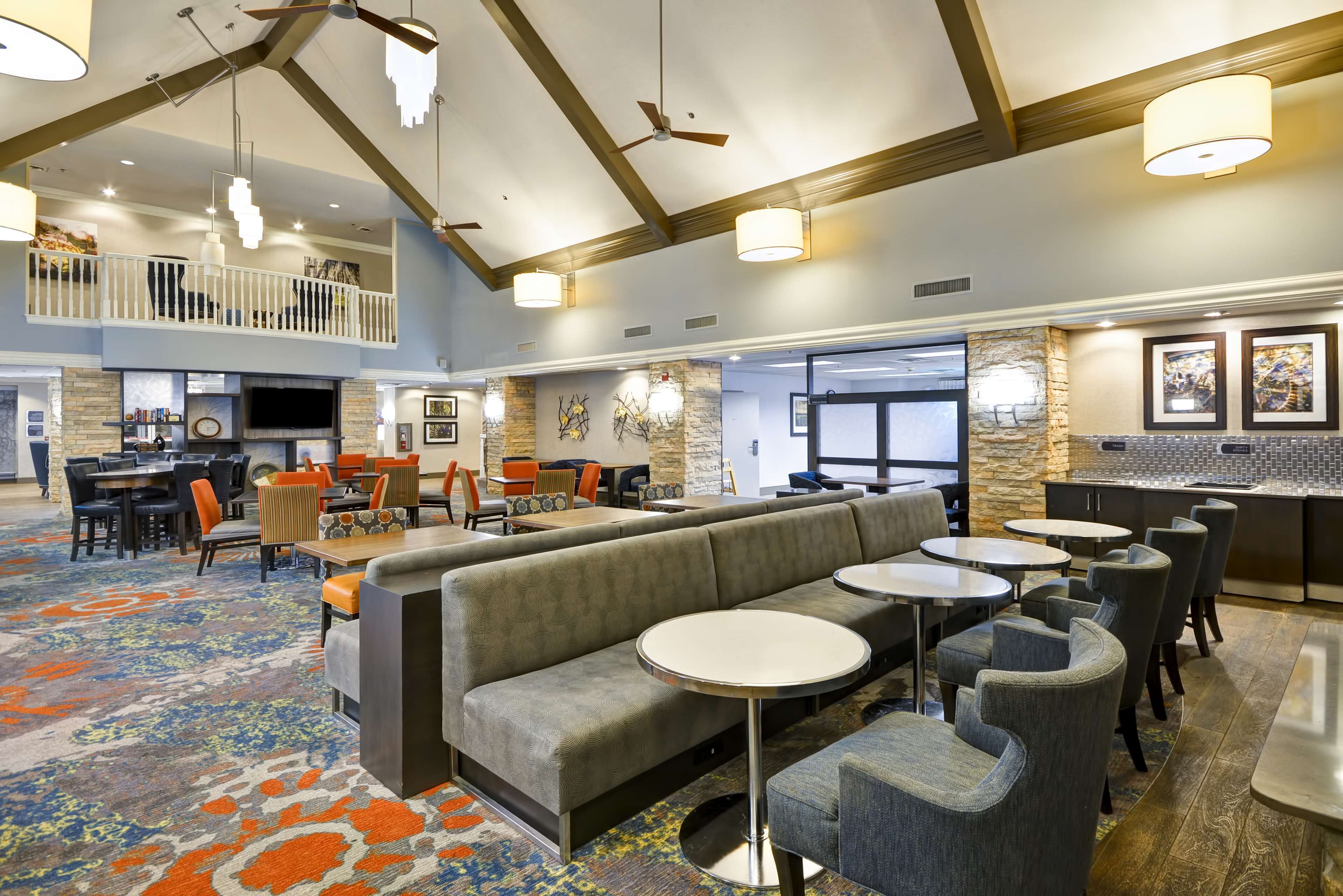 A spacious lobby with a high ceiling, a central seating area with a sofa and round tables, and a bar area with a stone accent wall.