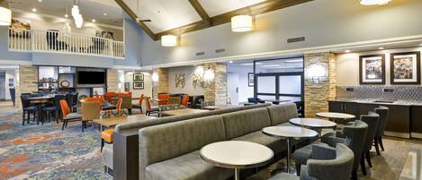 A spacious lobby with a high ceiling, a central seating area with a sofa and round tables, and a bar area with a stone accent wall.