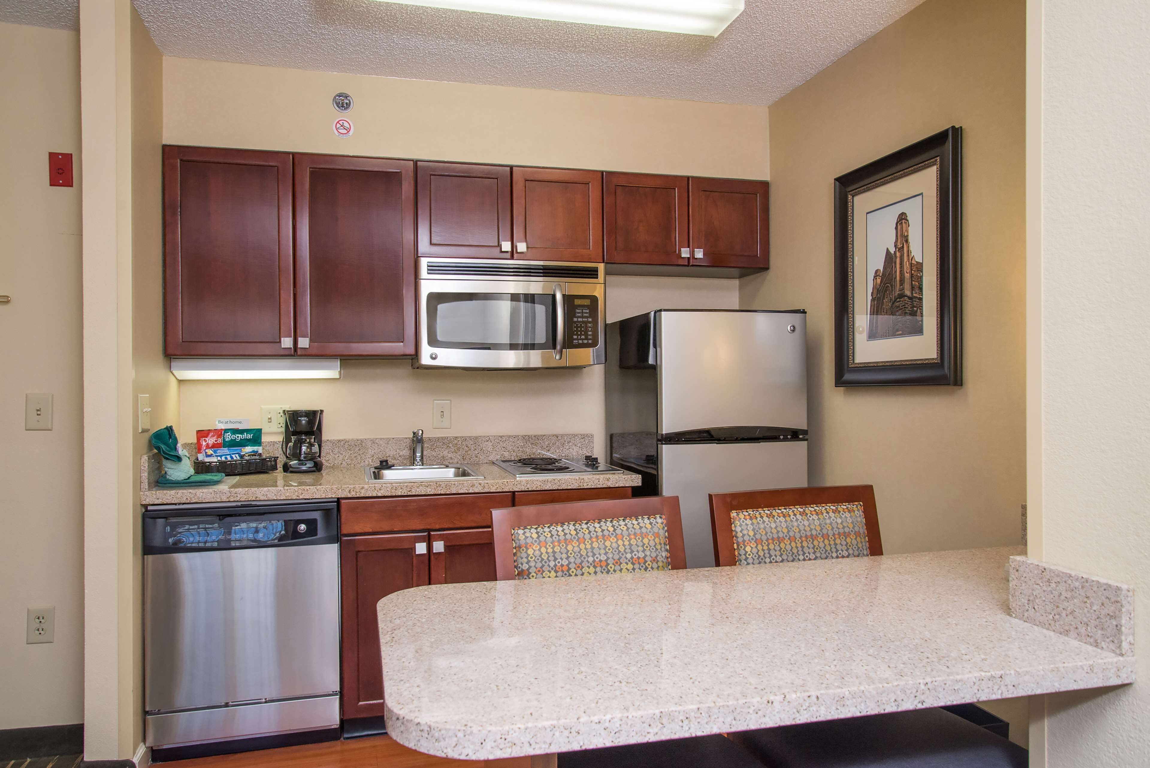 A compact kitchen with wooden cabinets, a stainless steel microwave, a refrigerator, and a dishwasher. There is a small island with a countertop and two chairs.