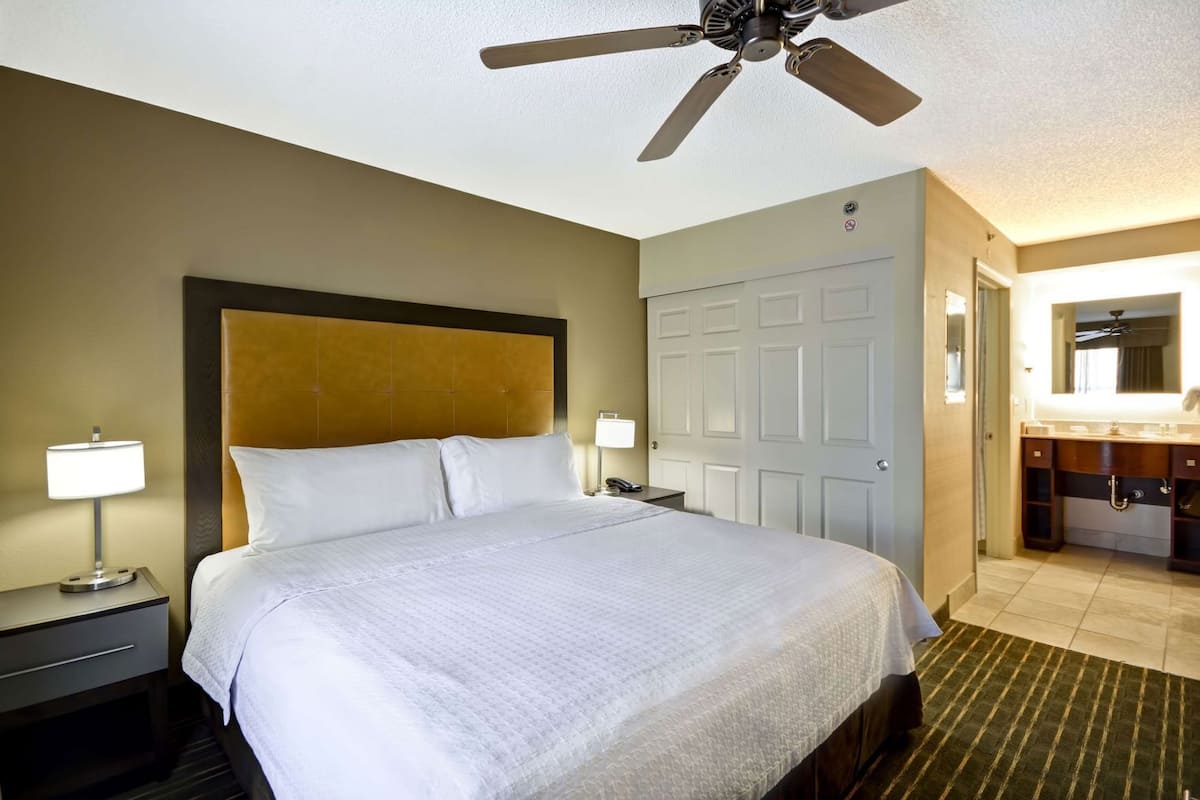 Habitación de hotel con cama, mesitas de noche, ventilador de techo, puerta y baño con lavamanos y espejo.