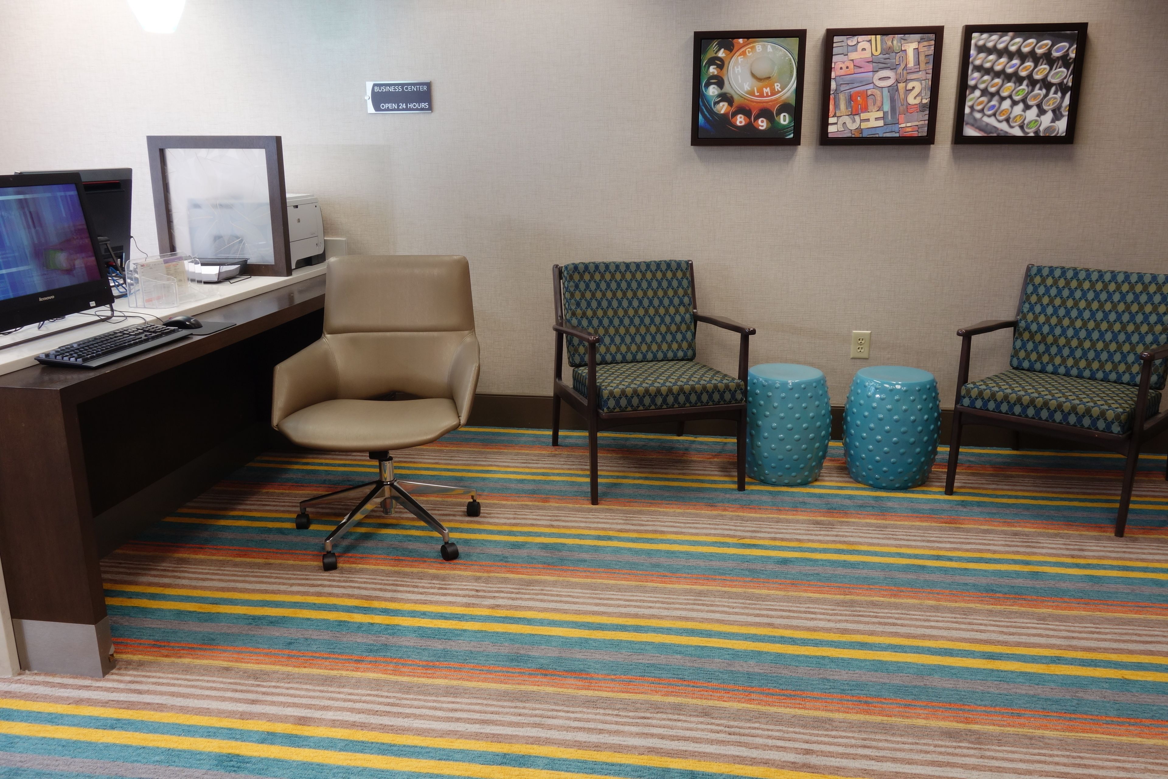 A hotel room with a desk, computer, two patterned chairs, and a colorful striped carpet.