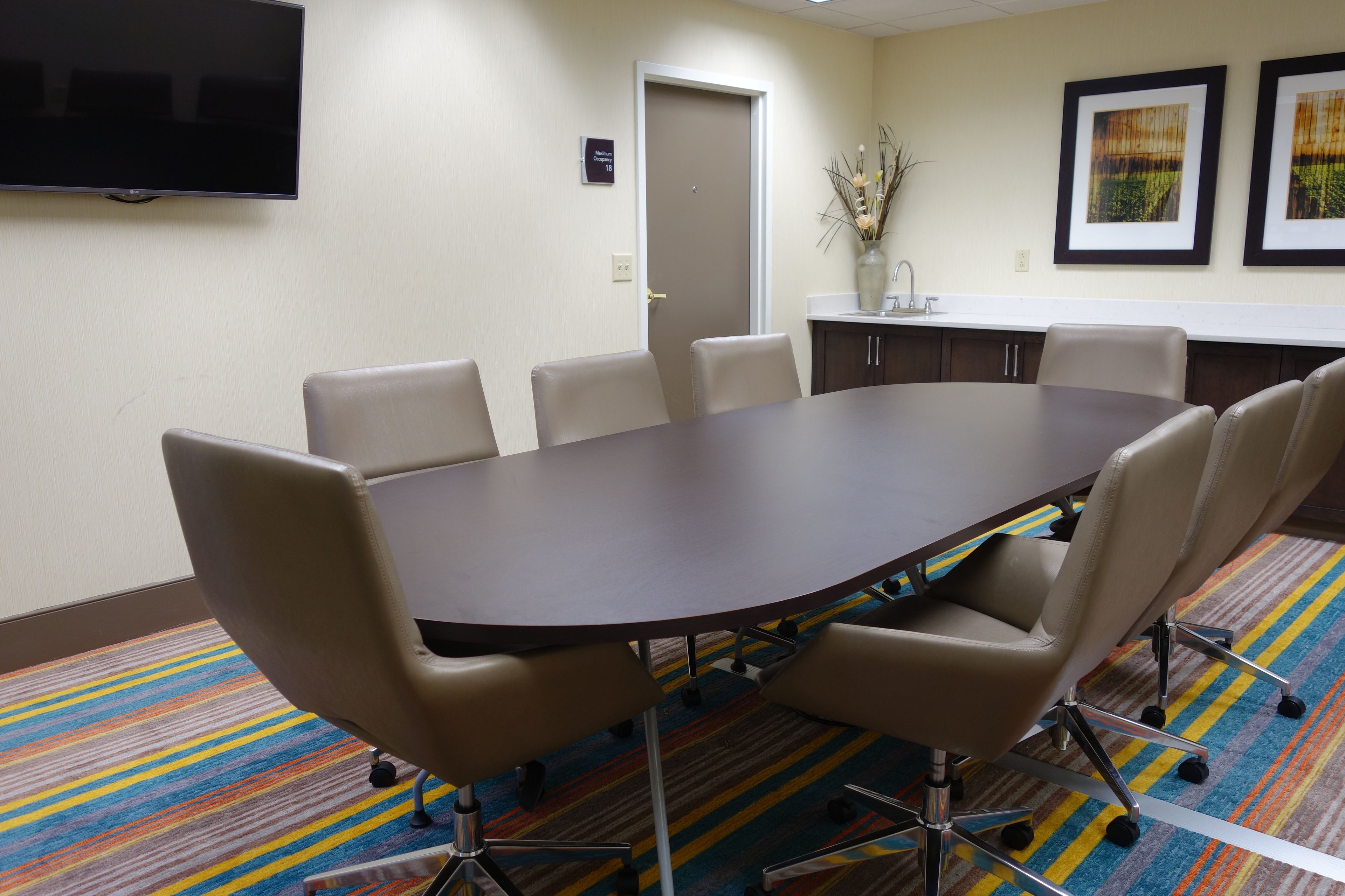 A conference room with a large table and six chairs, a flat-screen TV mounted on the wall, a door, and framed artwork on the wall.