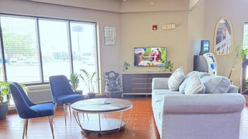 Un vestíbulo de hotel con una mesa redonda, sillas azules, un sofá gris, un televisor y un cajero automático.