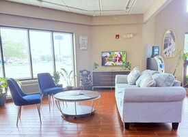 Un vestíbulo de hotel con una mesa redonda, sillas azules, un sofá gris, un televisor y un cajero automático.