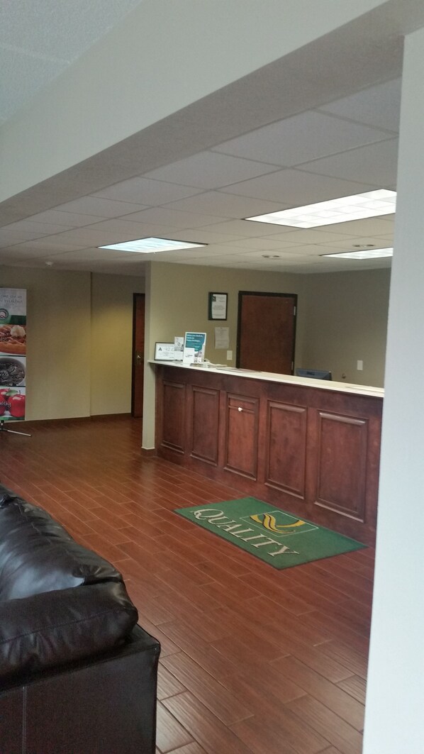 A reception area with a wooden counter, a green doormat with 'QUALITY' text, and a whiteboard on the wall.
