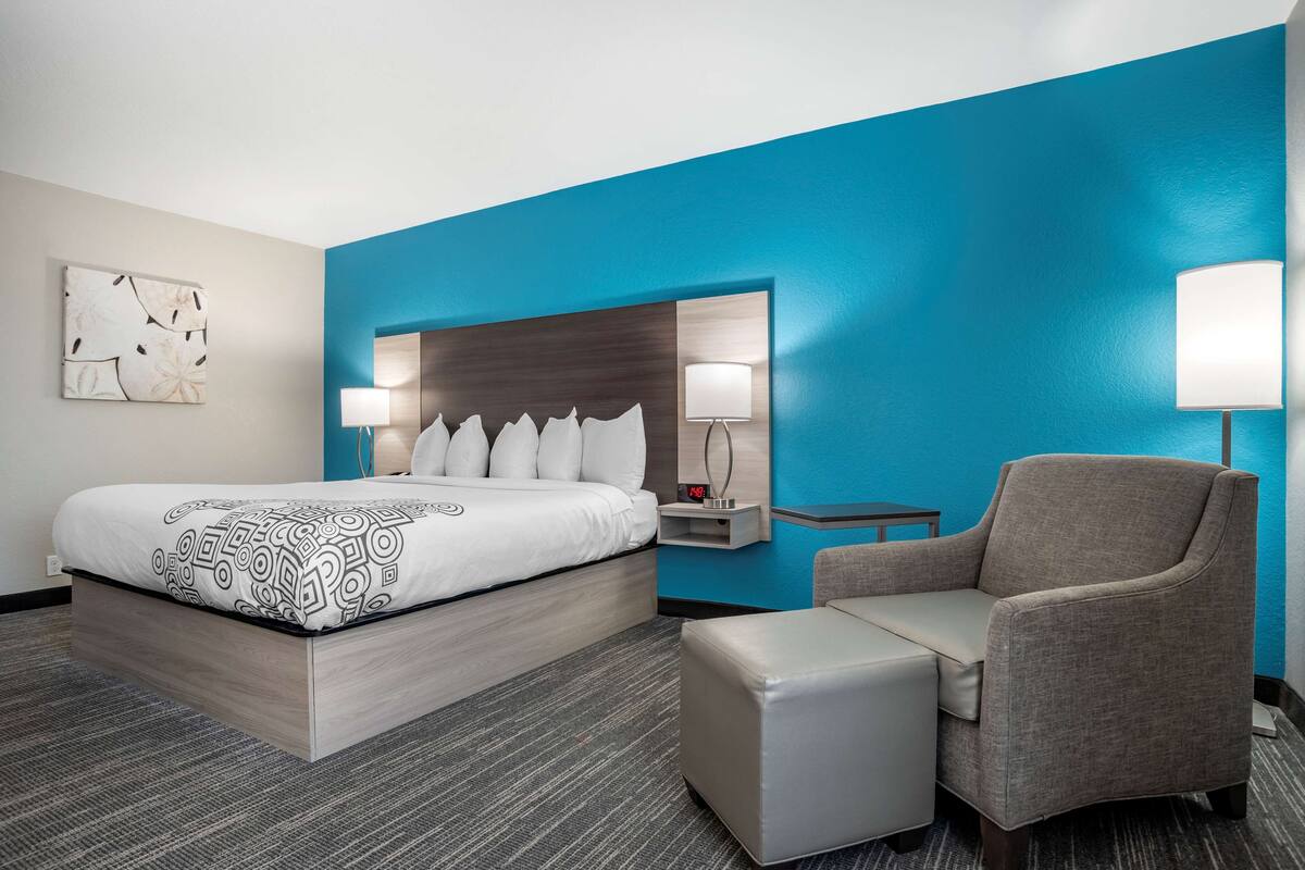 A hotel room with a large bed, a grey armchair, a small table, and a wall-mounted nightstand with a lamp.