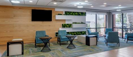 Un área de espera moderna con asientos cómodos, una televisión de pantalla plana y una pared verde con plantas.