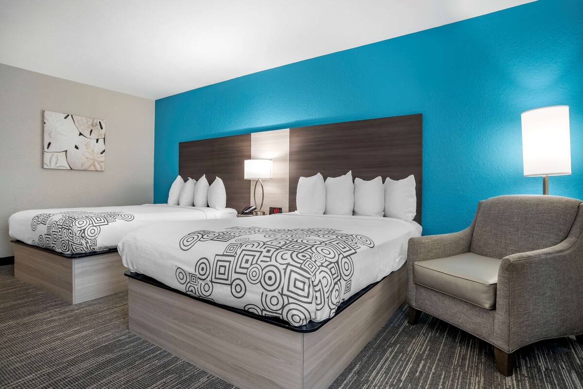 A hotel room with two beds, a chair, a nightstand, and a wall with a blue accent.