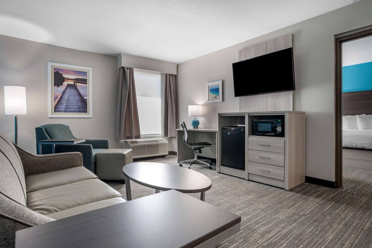 A hotel room with a sofa, a small table, a TV, a mini-fridge, and a desk with a lamp.