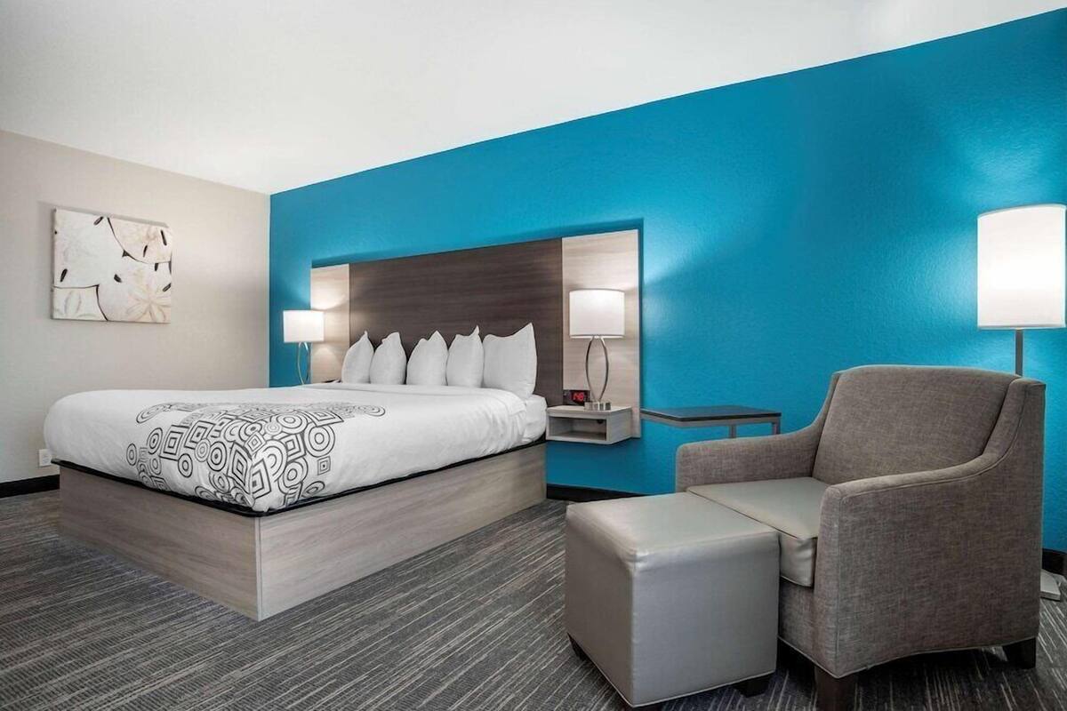 A hotel room with a bed, a chair, a nightstand, and a wall-mounted lamp.