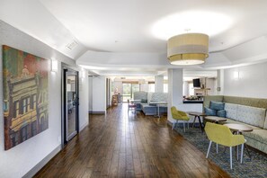 A modern hotel lobby with a wooden floor, a large painting on the left, a sofa and chairs in the center, and a kitchen area in the background. - Hampton Inn Shelbyville (Shelbyville)