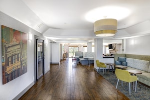 A modern hotel lobby with a wooden floor, a large painting on the left, a sofa and chairs in the center, and a kitchen area in the background.