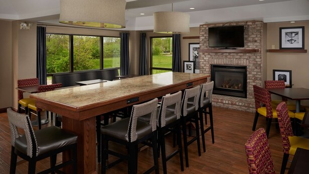 A spacious living area with a large wooden island, multiple seating options, a fireplace, and a television mounted on a stone wall. - Hampton Inn Shelbyville (Shelbyville)