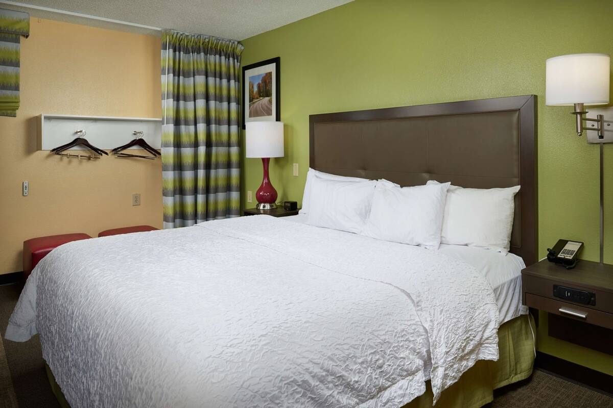 A hotel room with a large bed, a nightstand with a lamp, a red chair, and a wall-mounted coat rack.