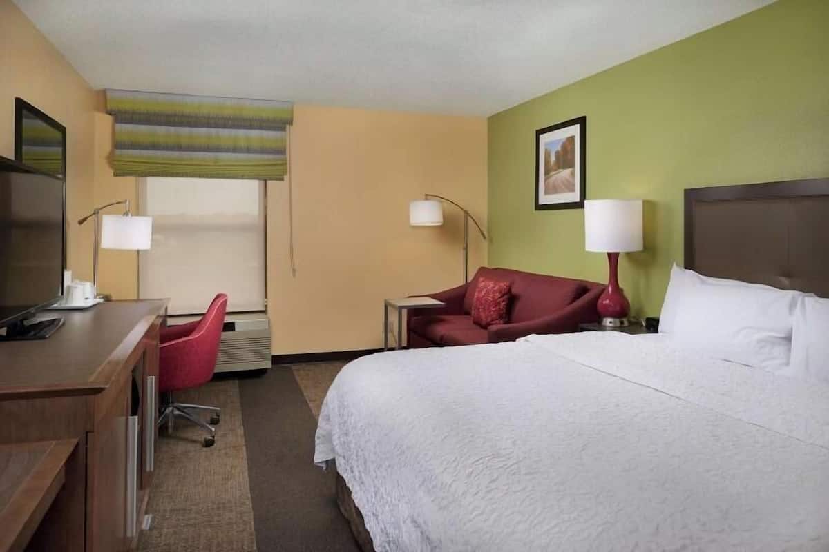A hotel room with a bed, a desk, a chair, a lamp, and a window with a view.
