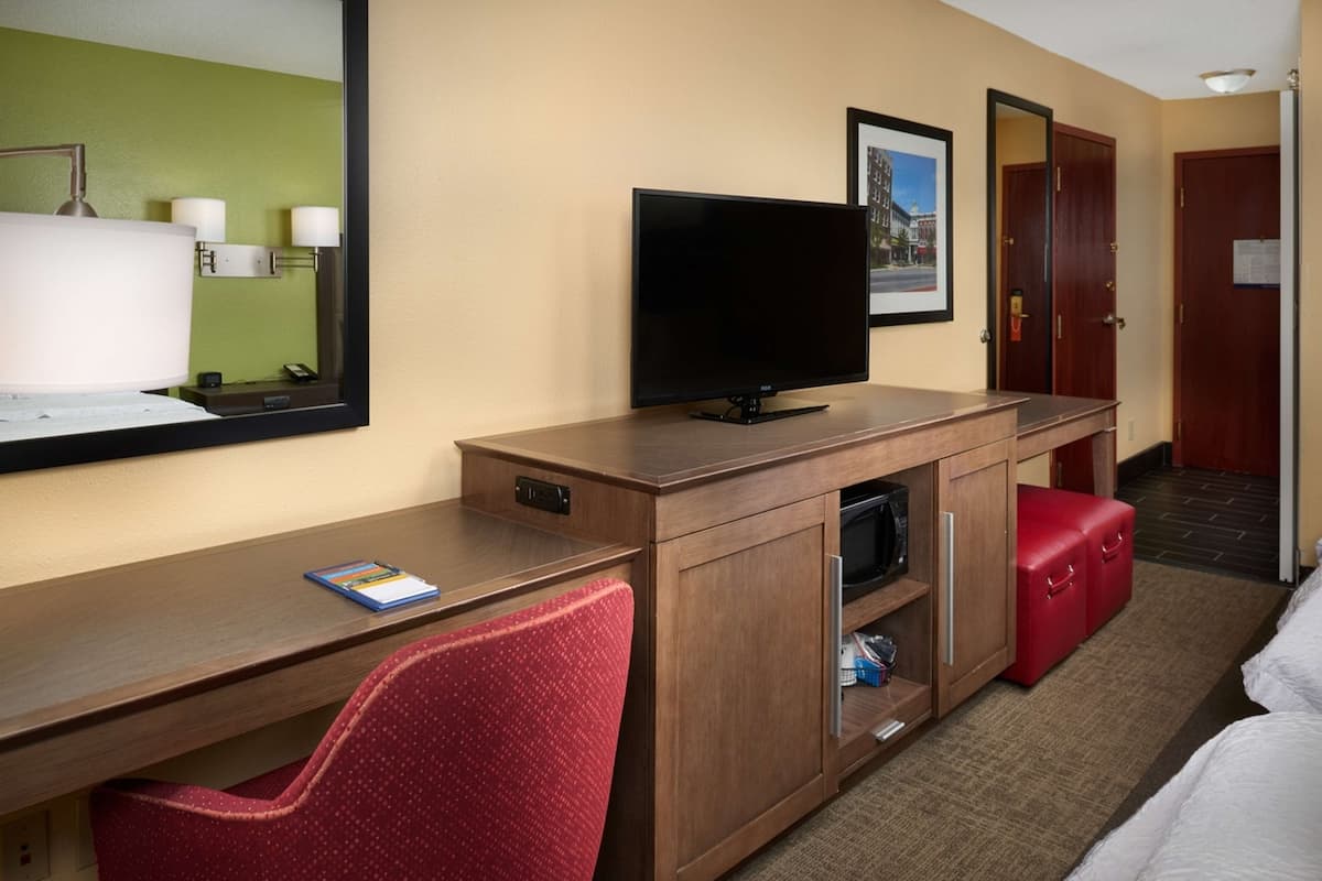 A hotel room with a wooden desk, a television, a red chair, a red storage bench, and a large mirror.