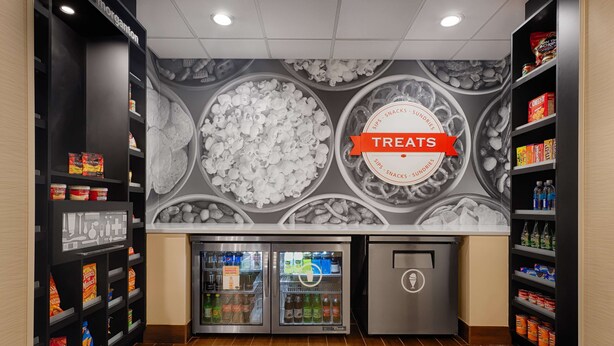 A snack bar with shelves stocked with various snacks and drinks, a 'Treats' sign, and a refrigerator. - Hampton Inn Morganton (Morganton)