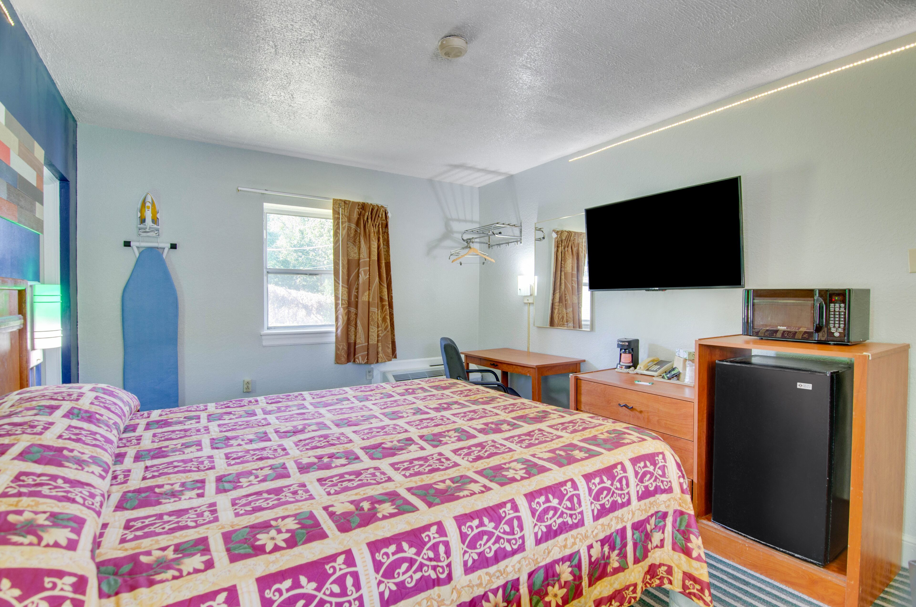 A hotel room with a large bed, a desk with a chair, a television, a refrigerator, and a window with curtains.