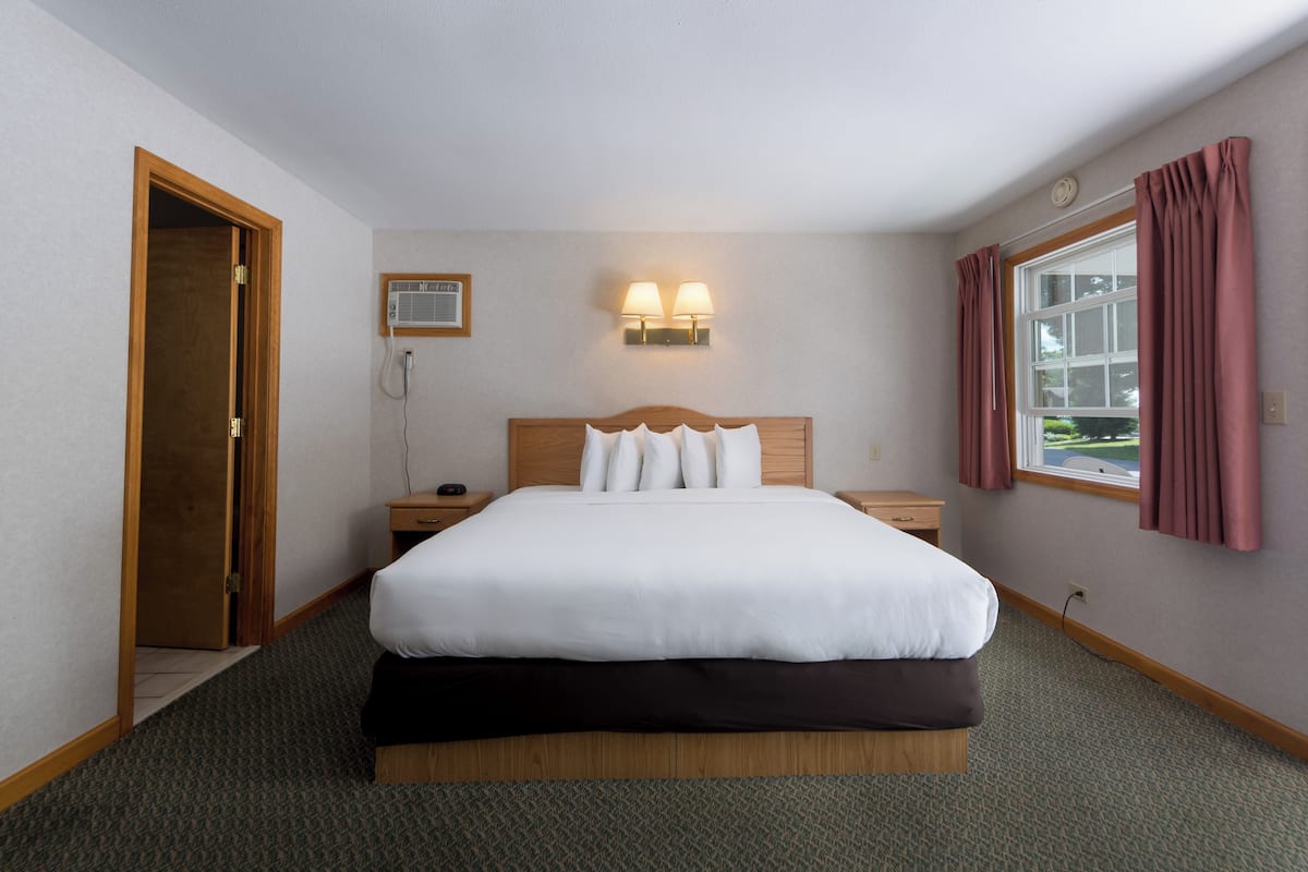 A hotel room with a large bed, wooden headboard, two bedside lamps, a window with curtains, and a door leading to another room.