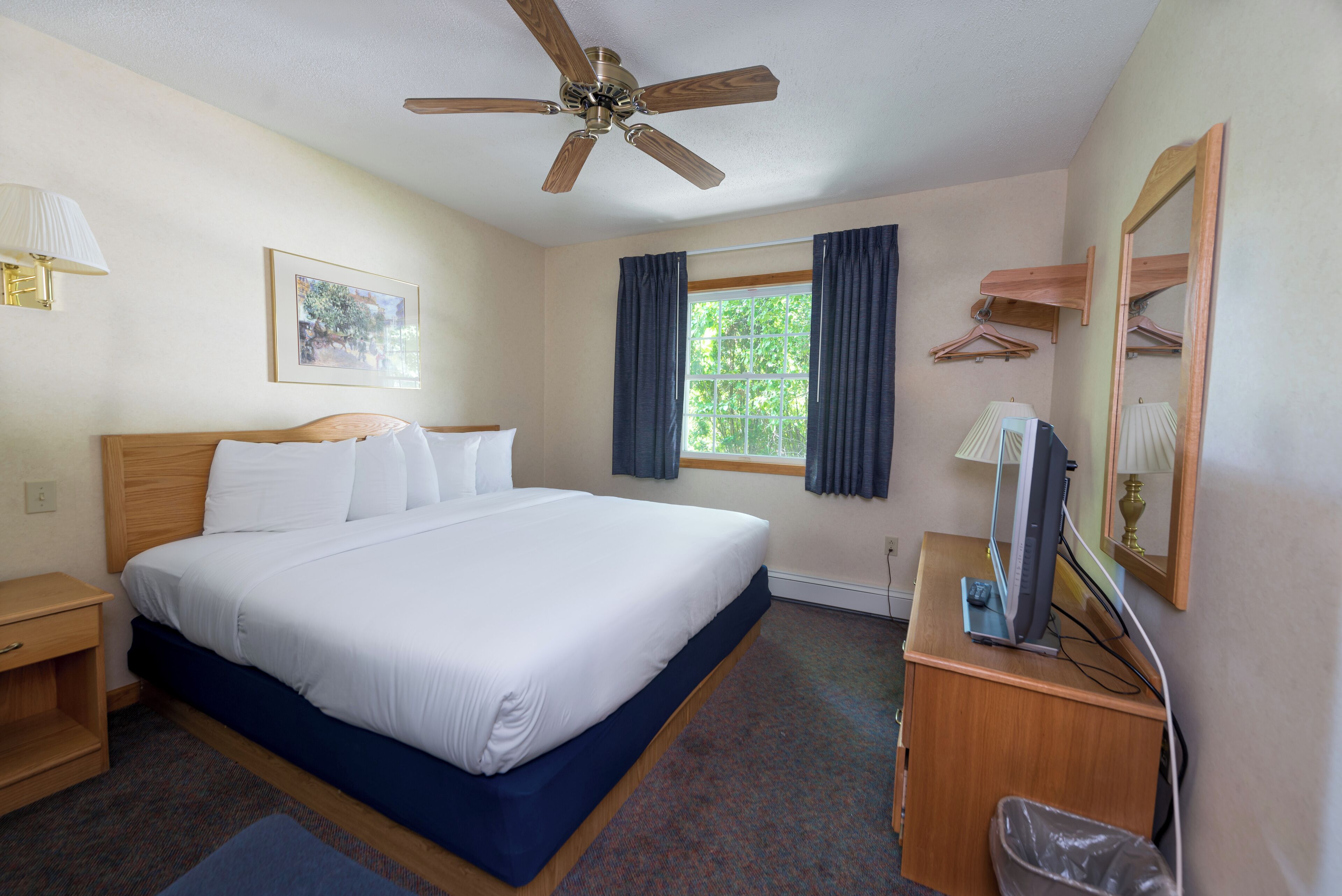 A hotel room with a bed, a desk with a computer, a ceiling fan, a window with curtains, a lamp, and a framed picture on the wall.