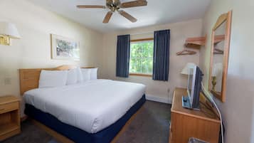 Habitación de hotel con cama, escritorio con computadora, ventilador de techo, ventana con cortinas, lámpara y un cuadro en la pared.