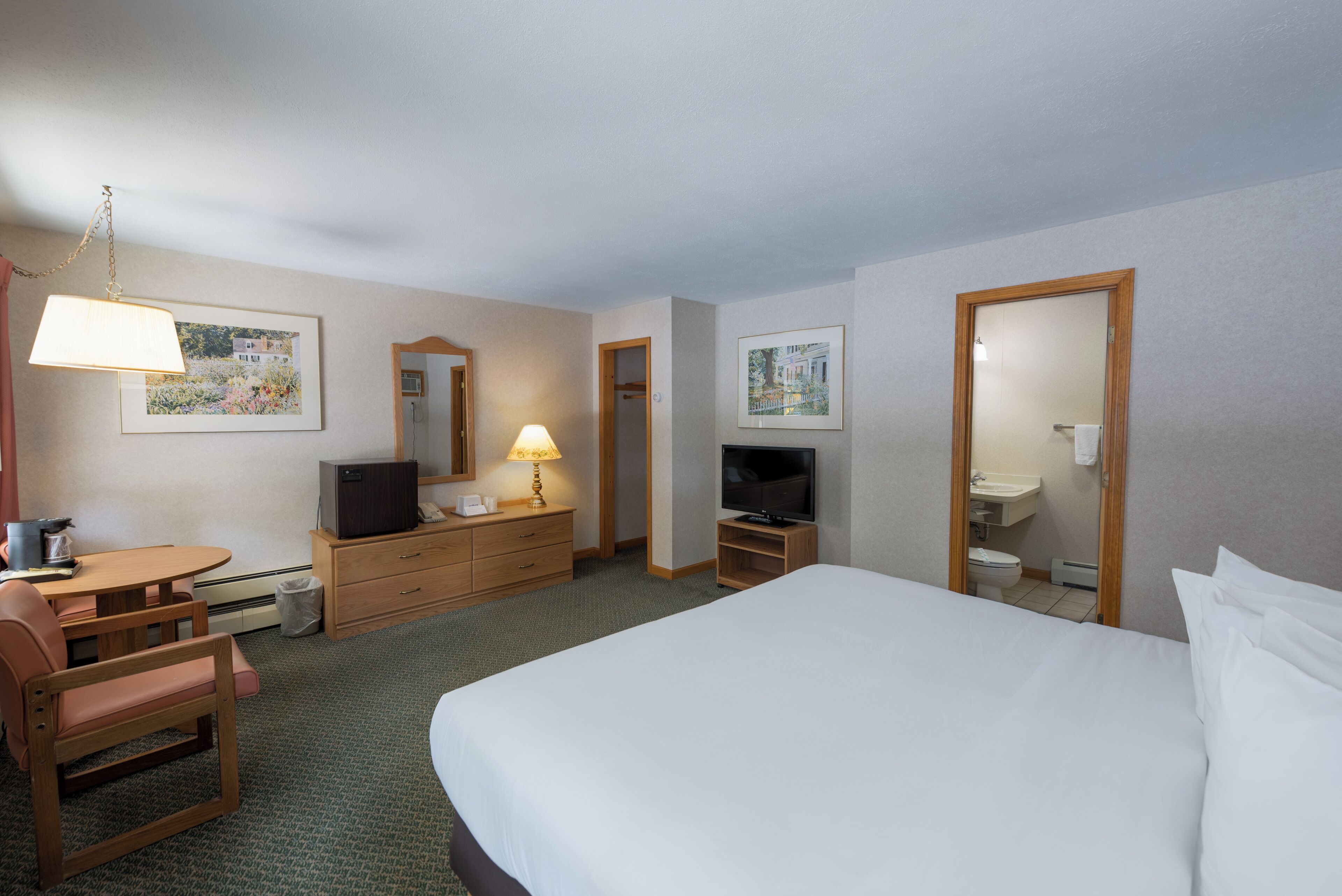 A hotel room with a bed, a desk with a chair, a television, a dresser, and a bathroom visible through an open door.